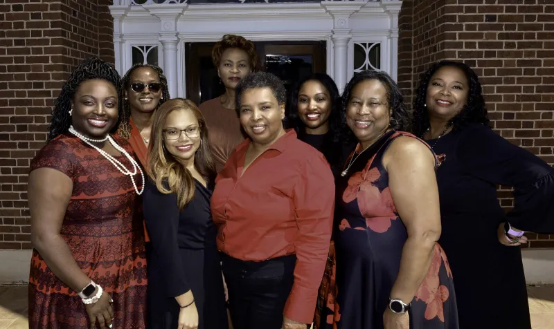 A group of eight women smiling in front of a brick building.