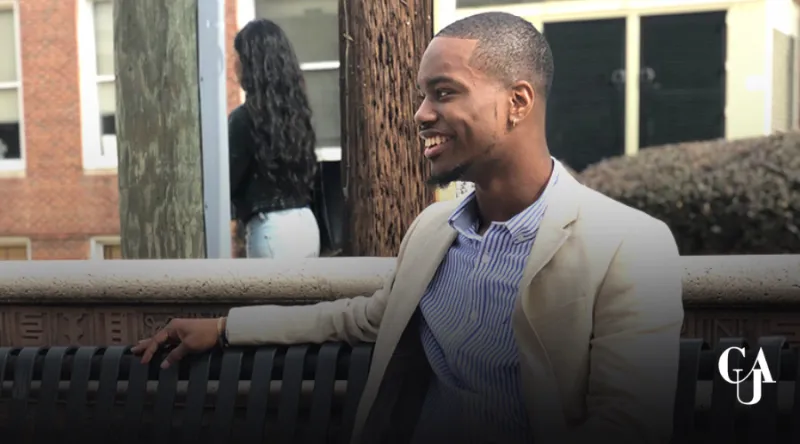 Man in a light jacket smiles on a bench, woman walks in the background.