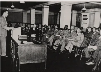 A man speaks to a crowd seated in a dimly lit room.