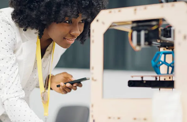 Woman observing a 3D printer model with curiosity, holding a phone.