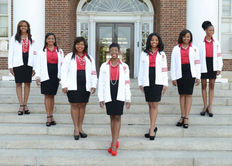 Seven women in matching white jackets and red tops stand on building steps, smiling.