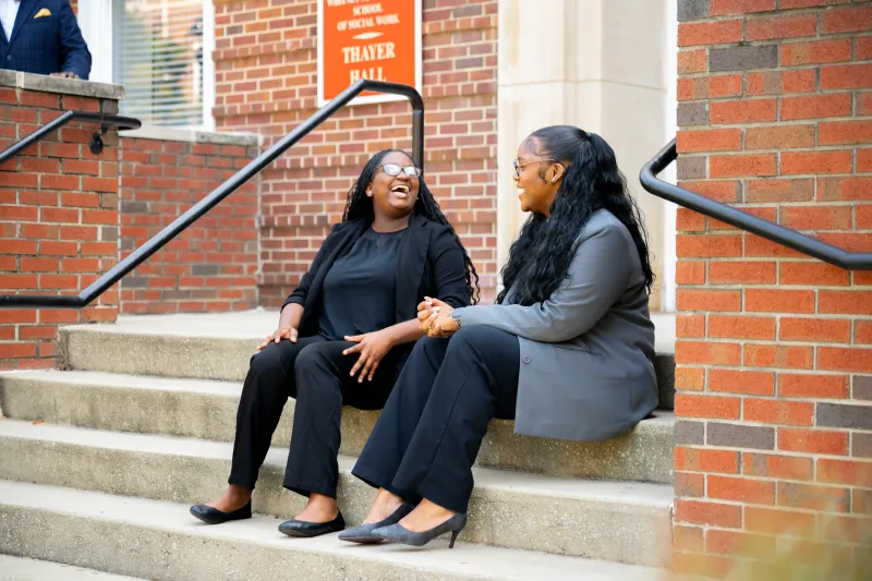 Two students in front of WMYJ SSW in business attire laughing on brick building steps.