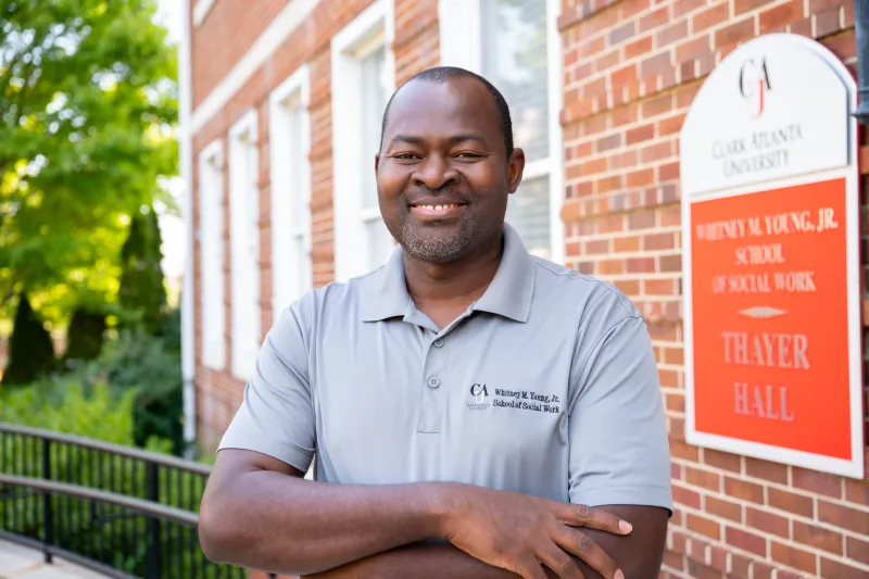 A man smiles, standing outside a brick building with a white and red sign.
