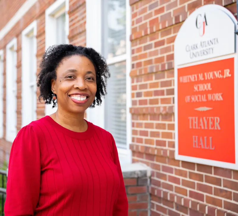 Smiling woman in red sweater outside brick building with university sign.