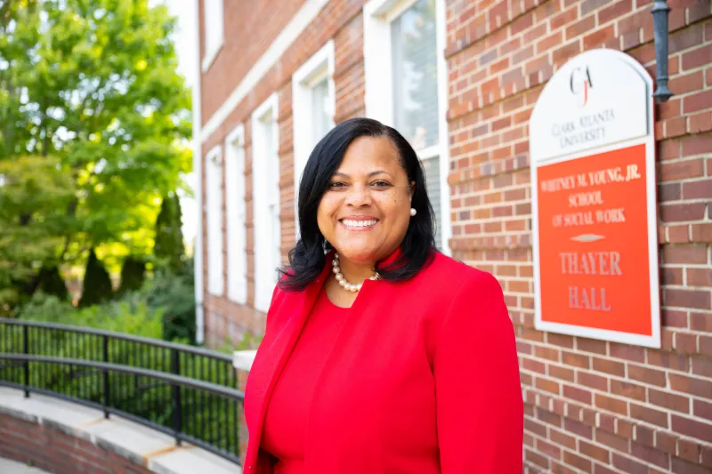 Dean S. Jackson in red suit smiling outdoors near a brick building and sign.