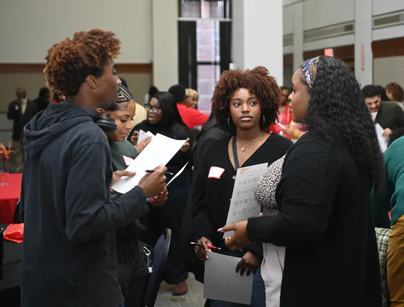 People in a group discussion at an event, holding papers, with a busy background.