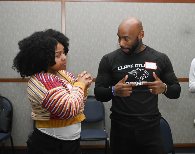 Two people talk in a room, one wears a striped sweater, the other a black shirt.