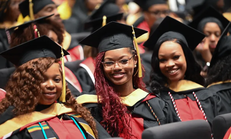 Graduates in caps and gowns smiling, seated in a crowded ceremony.