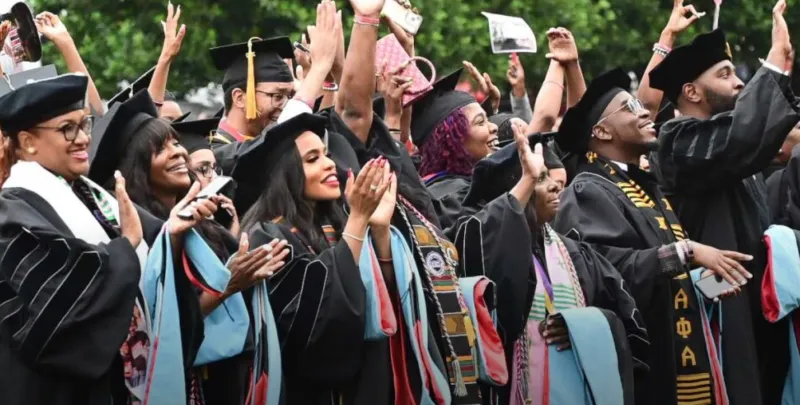 Graduates celebrating in caps and gowns, smiling and clapping outdoors.