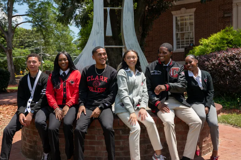Six students sitting on a brick wall, smiling on a sunny day.
