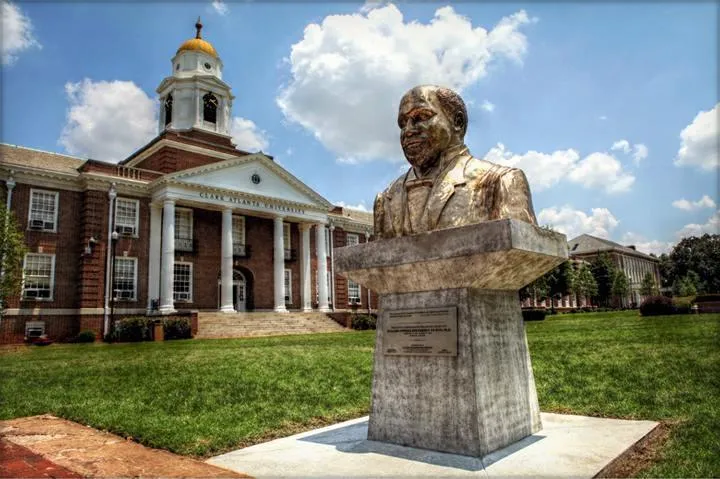 Bust sculpture on campus lawn, large brick building behind, sunny sky with clouds.