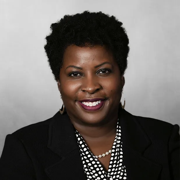 Smiling person in a black blazer and polka dot blouse against a grey backdrop.
