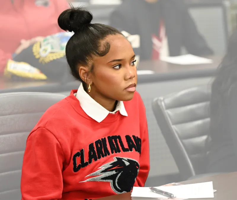Young woman in red sweatshirt sits attentively in a classroom.