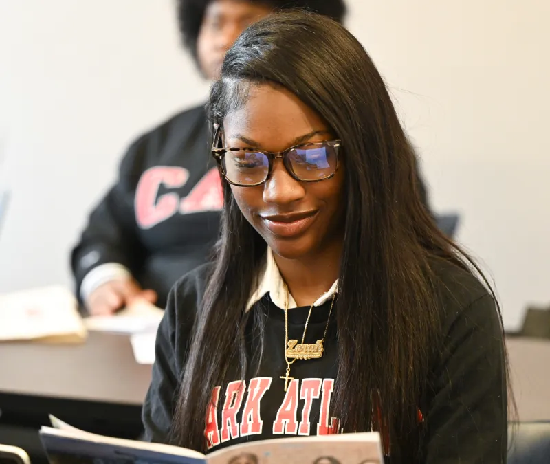 Student at desk reading a book, wearing glasses and a black sweater.