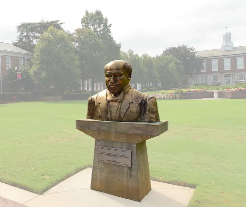 Bronze bust of a person on a pedestal in a park with trees and a building in the background.