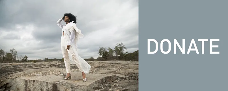 Person in white standing on a rocky landscape under a cloudy sky, text says “DONATE.”