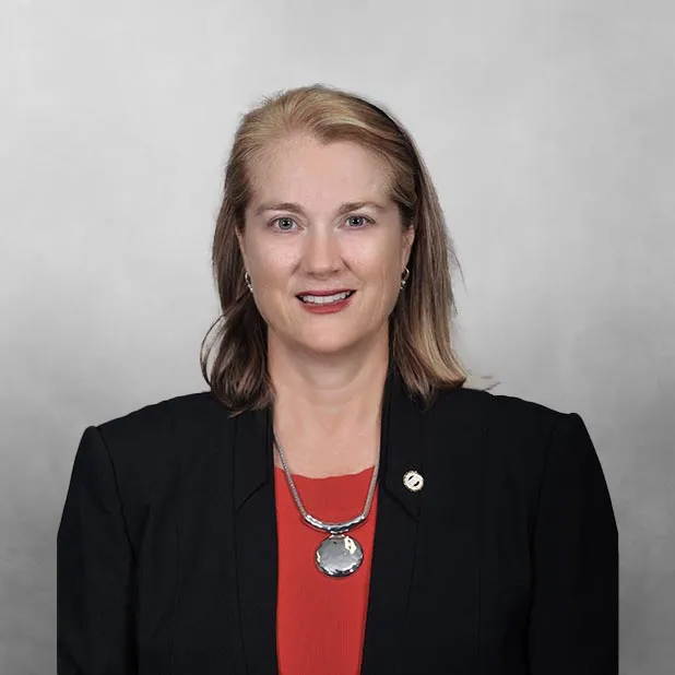 Professional woman in black blazer and red top against gray background.
