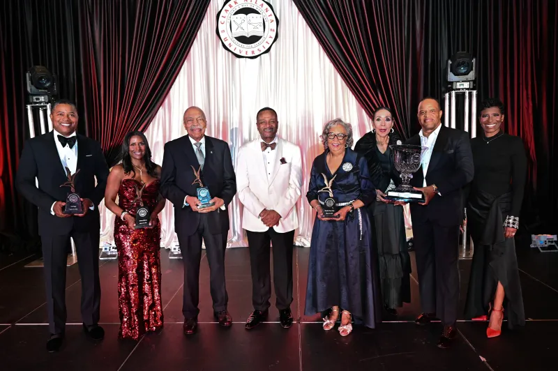 A group of people in formal attire holding awards on stage with curtains in the background.