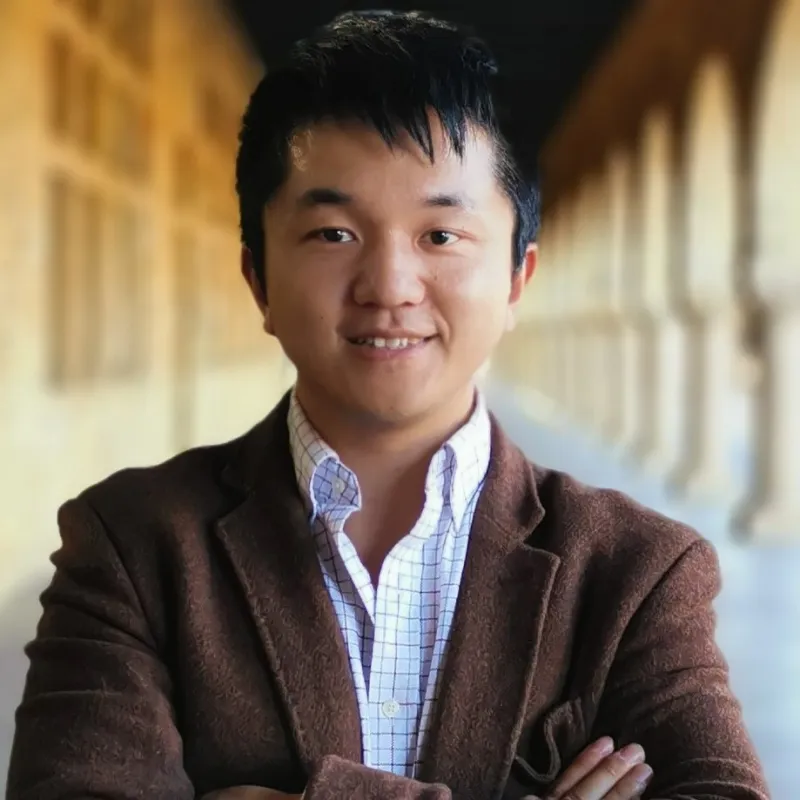 Man in a brown jacket smiling, standing in a corridor.