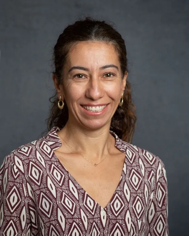 Smiling woman with curly hair, wearing a patterned shirt against a gray background.
