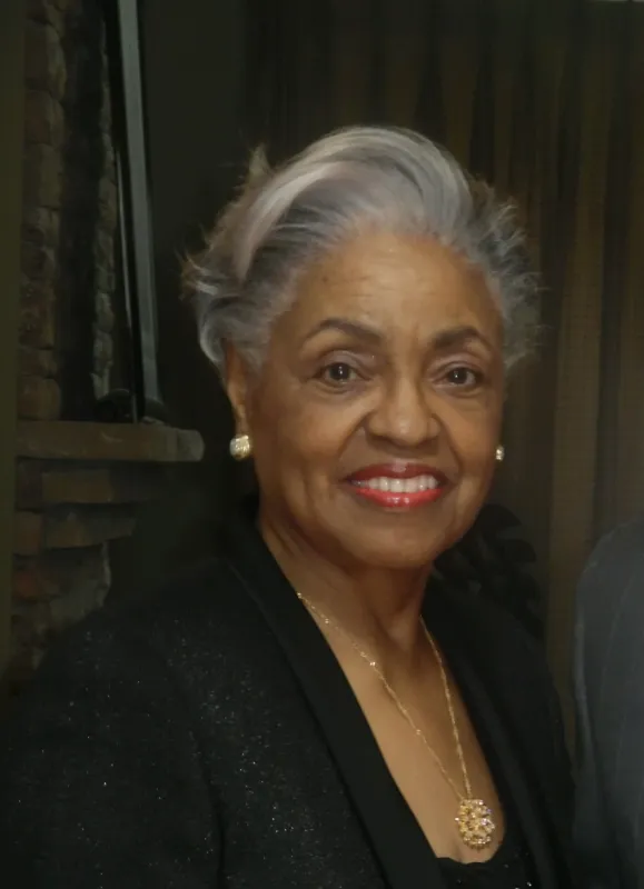 Elegant older woman with gray hair, smiling in a black outfit and pearl earrings.