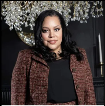 Woman in a brown suit poses confidently under a crystal chandelier.