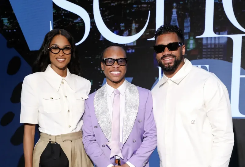 Three people smiling in front of a backdrop, wearing formal attire and sunglasses.