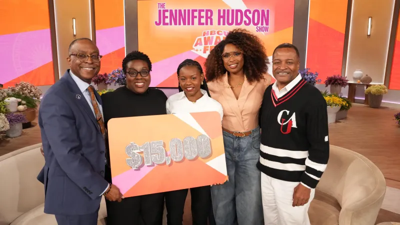 Five people smiling on a talk show set, holding a $15,000 sign.