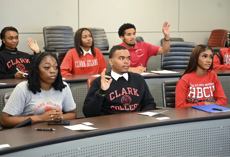 Students in a classroom, some raising hands, wearing college sweatshirts.