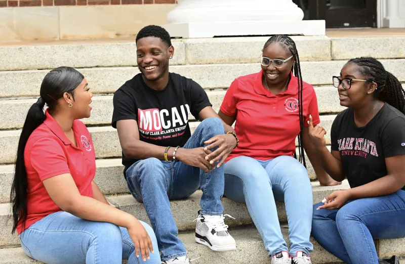 Four students sitting on steps, smiling and talking, wearing casual clothes.