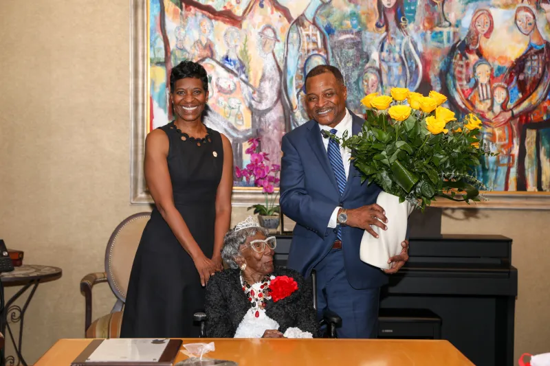 Smiling trio in front of colorful artwork; man holding yellow flowers.