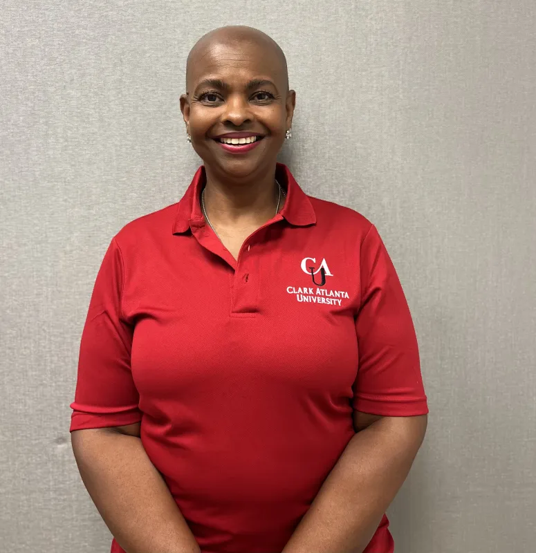 Smiling person in a red polo shirt with a logo, standing against a gray wall.